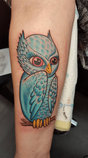 Ein farbenfrohes Tattoo einer blauen Comic-Eule mit großen rosa Augen auf dem Arm. Eulen-Tattoo von Ozzy in Hamburg gestochen.