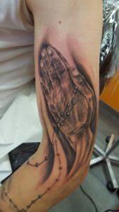Realistische betende Hände mit einem Rosenkranz als detailreiches Black and Grey Tattoo auf dem Oberarm. Dürers-betende-Hände-Tattoo von Ozzy in Hamburg gestochen.
