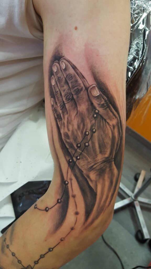 Realistische betende Hände mit einem Rosenkranz als detailreiches Black and Grey Tattoo auf dem Oberarm. Dürers-betende-Hände-Tattoo von Ozzy in Hamburg gestochen.