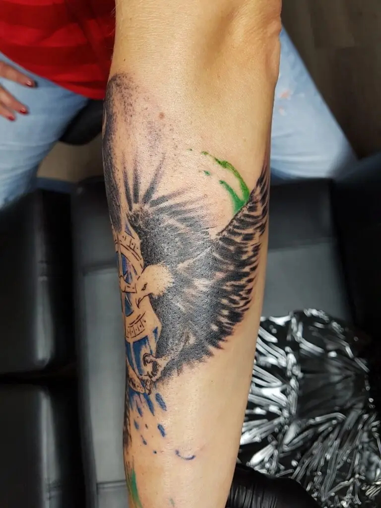 Weißkopfseeadler-Tattoo auf dem Unterarm. Es zeigt einen Adler mit einem blauen Schild als Teil eines Amerika-Unterarm-Sleeves. Adler-Tattoo gestochen von Ozzy in Hamburg Black-and-Grey mit blauen und grünen Farbspritzern. Gestochen von Ozzy in Hamburg.