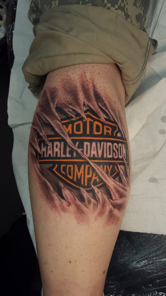 Das Harley-Davidson-Logo, das durch aufgerissene Haut auf der Wade sichtbar wird. 3D-Tattoo von Ozzy in Hamburg gestochen.