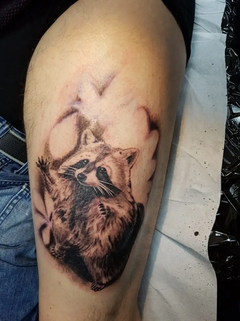 Realistisches Black-and-Grey Tattoo eines süßen, winkenden Waschbären auf einer Wade, gestochen von Ozzy in Hamburg.