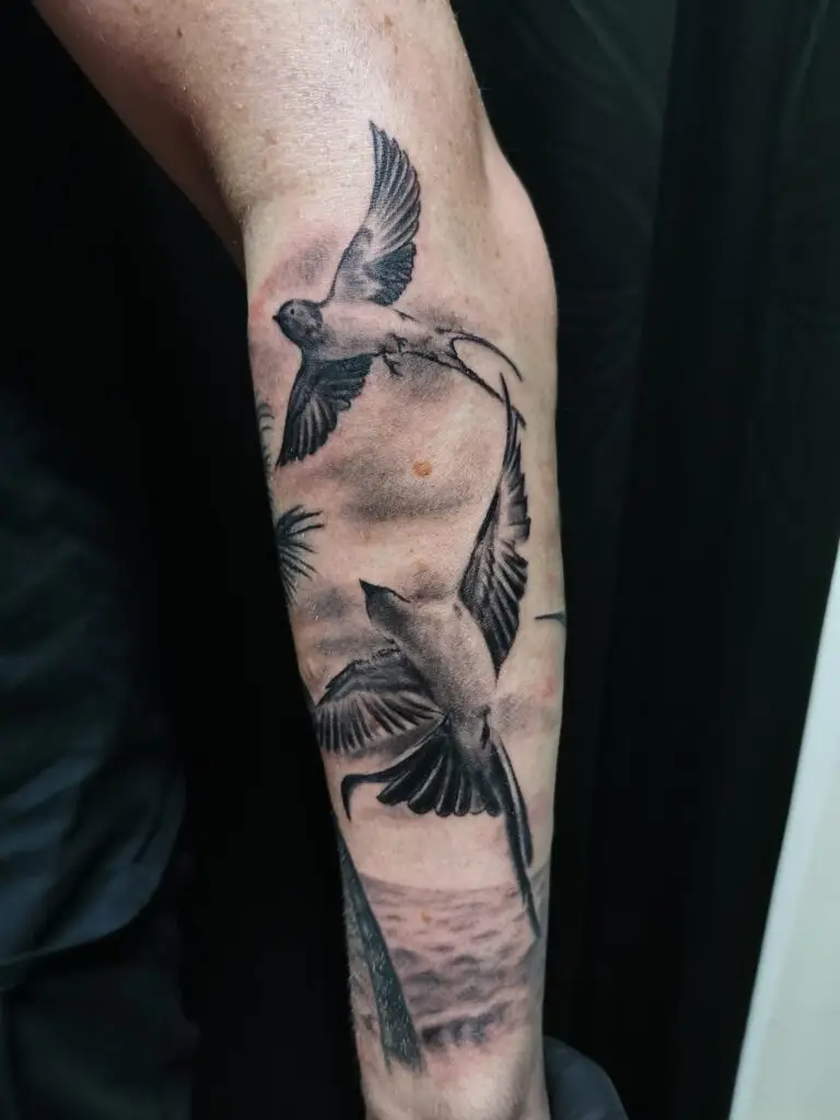 Ein Black-and-Grey-Tattoo auf dem Unterarm, das zwei fliegende Schwalben über einer Strandszene mit Palmen zeigt — Schwalben-Tattoo von Ozzy in Hamburg gestochen.