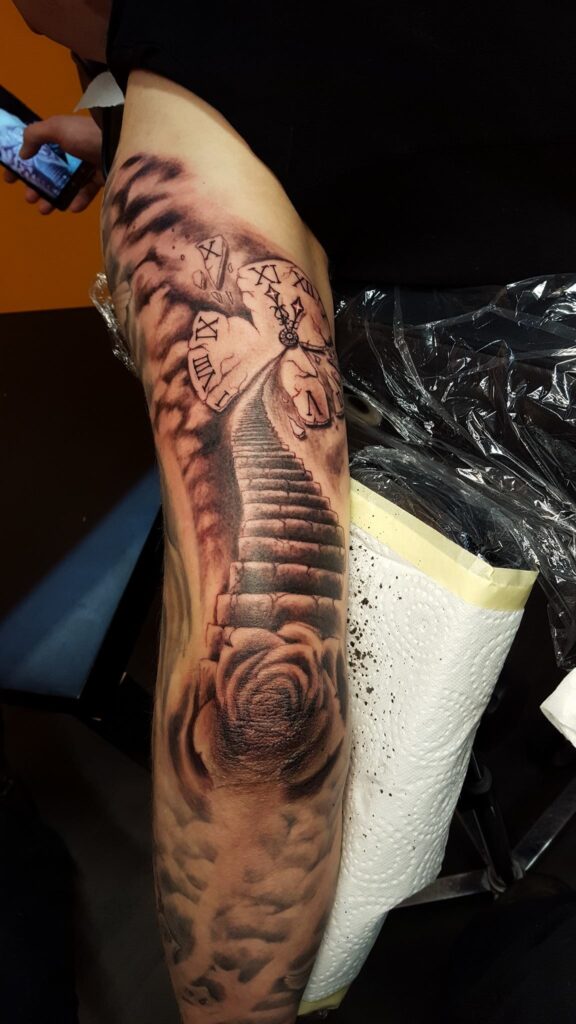 Ein surreales Tattoo einer Treppe, die zu einer Uhr im Himmel führt, mit einer Rose am Anfang. Surreales-Tattoo von Ozzy in Hamburg gestochen.